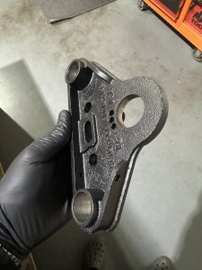 A gloved hand holds a black, metal automotive part with cylindrical openings and various markings, against a gray workshop floor.