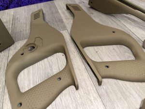 Two tan, textured plastic or polymer handle components with cutouts, resting on a light wood surface.