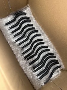 A cardboard box containing fifteen identical black metal rods with silver ends, arranged in a row on bubble wrap.