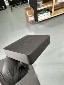 A gloved hand holds a sleek, black rectangular device with a logo engraved on top; shelves and a cardboard box are visible in the background.