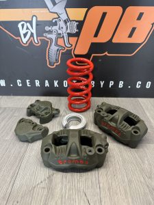 Olive green Brembo brake calipers, a red coil spring, and a silver metal ring displayed on a wooden surface with a branded backdrop.