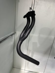 Matte black, curved metal pipe with two branches, hanging from a hook inside a light-colored enclosure.