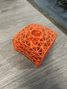 A 3D-printed orange geometric structure with a lattice design and a circular hole in the center sits on a wood-grain surface.