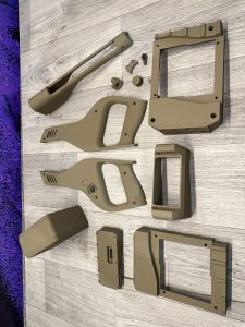 Several tan-colored plastic or metal device casing parts are arranged on a light wood floor. The components vary in size and shape, suggesting disassembled equipment housing.