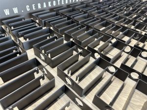 Rows of identical black metal brackets with circular cutouts lined up on a wooden surface, with a Cerakote branding sign in the background.