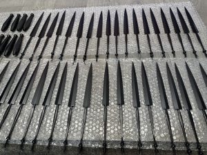 Several rows of black metal knives with handles are arranged neatly on sheets of bubble wrap on a wooden floor.