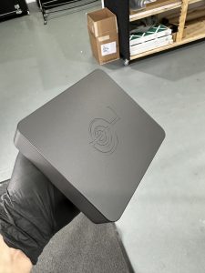 A hand wearing a black glove holds a matte black rectangular box with a circular geometric logo on the lid; shelves and cardboard boxes are visible in the background.
