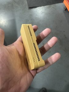 A hand holding a small, rectangular, gold-colored metal object with vent-like slots on one side and rounded edges.