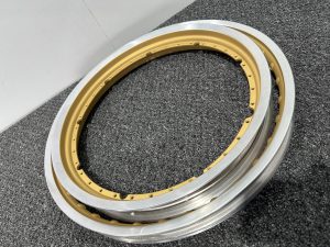 Two metallic circular wheel rims, one gold and one silver, are placed on a gray textured surface against a white wall.