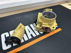 Two gold-colored mechanical parts, possibly automotive components, are placed on a black mat with white and orange text.