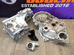 Aluminum automotive transmission case components and brackets displayed on a wooden floor, with a branded backdrop in the background.