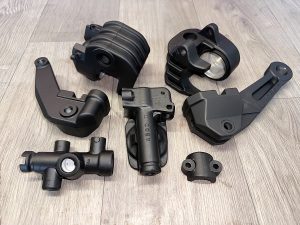 Seven black metal automotive parts arranged on a light wood surface, each with distinct shapes, holes, and mounting points.