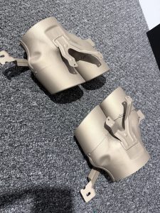 Two matte beige metal exhaust tips with mounting brackets are placed on a textured gray carpet.